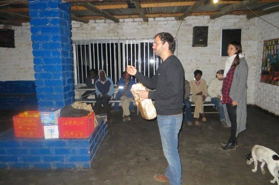 Pastor Wenzel speaking to men seated on benches at night, with Sharon beside him